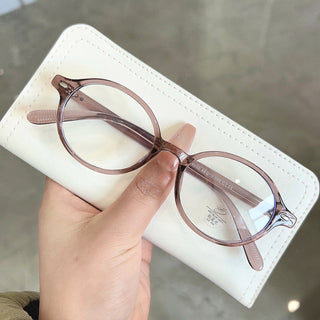A hand with long, nude nails holds EyeCandys Chase Round Oval glasses and a white wallet against a gray background.