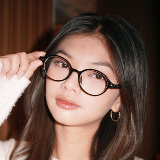 A young woman with long, straight brown hair wears EyeCandys Chase Round Oval glasses and gold hoop earrings. She slightly smiles, holding her glasses with one hand and looking directly at the camera.