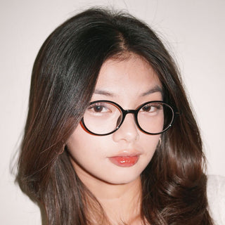 A person with long dark hair wears EyeCandys Chase Round Oval eyeglasses, looking at the camera with a neutral expression against a plain light background.