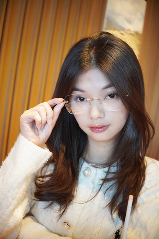 A woman with long dark hair wears a white sweater and holds her EyeCandys Dusk Rimless Rectangular eyewear, looking at the camera indoors against a blurred wooden background.