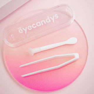 A transparent plastic case branded with "EyeCandys" is open, displaying a white tweezer-like tool and an applicator on a pink and white gradient surface. This EyeCandys Contact Lens Applicator Kit (GWP) appears to be designed for handling contact lenses with ease and precision.