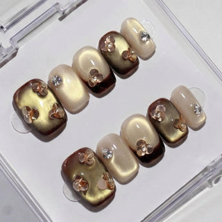 EyeCandys Gilt Trip Press-On Nails are handmade with a glossy finish in beige, gold, and brown. They feature cat-eye gold accents, rhinestones, translucent floral designs, and come neatly arranged in a clear plastic case.