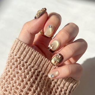 A hand wears EyeCandys Gilt Trip Press-On Nails, gold and brown with rhinestones and pearl accents, partially covered by a beige knit sweater sleeve in natural light.