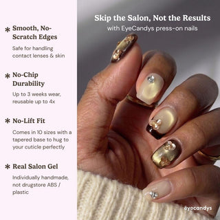 A hand shows off EyeCandys Gilt Trip Press-On Nails—long, almond-shaped in gold, brown, and nude with pearls and gold accents. Enjoy smooth edges, no-chip durability, a secure fit, and a real salon gel manicure finish.