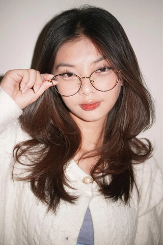 A woman with long dark hair wears EyeCandys Madame Cat Eye lightweight metal frames and a white textured top, looking at the camera while holding her glasses with one hand and slightly smiling.