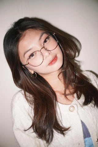 A young woman with long brown hair models EyeCandys Madame Cat Eye frames—lightweight metal in a subtle cat-eye style—while wearing a white textured sweater and light blue top against a plain light background.