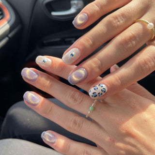 Hands with EyeCandys Purr-ple Reign Press-On Nails show iridescent purple nails, a blue gem accent, and a white base with black and blue leopard print. This stylish look highlights easy salon-quality nails and delicate rings.