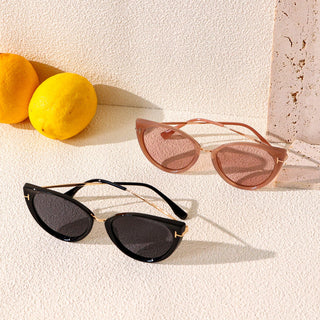 Two pairs of EyeCandys Vintage Cat Eye Sunglasses are displayed on a textured surface with two citrus fruits in the background. Shown are sunglass frames in black and in light pink, with gold accents.
