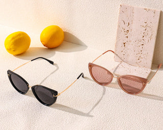 Two pairs of EyeCandys Vintage Cat Eye Sunglasses are displayed on a beige textured surface with two citrus fruits in the background. Shown are sunglass frames in black and in light pink, with gold accents.