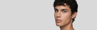 A man with short curly dark hair wearing light gray colored contacts against a plain background.