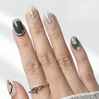 Hand wearing EyeCandys Concrete Poetry press on nails featuring cream and charcoal abstract nails with gold foil and ink wash detailing.