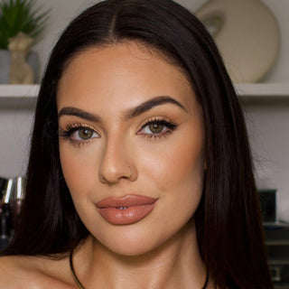 A woman with long, straight dark hair and clear, grey eyes, accentuated by EyeCandys Isla Mirage Grey prescription contact lenses from EyeCandys, is looking at the camera. She has flawless makeup, a nose ring, and is holding her right hand to her neck. The background appears to be a blurred room with some decor and objects.