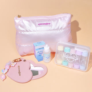 A pastel pink EyeCandys travel kit featuring a shiny puffy makeup pouch, a blue packaging box and bottle of Dream Eye lubricant eye drops, a clear case with colorful contact lens containers, and a pink heart-shaped mirror keychain with charms.