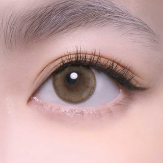 Close-up of a human eye wearing Bausch & Lomb Lacelle Merry Mocha (30pk) contact lenses, with long lashes, a well-groomed eyebrow, clear skin, and subtle makeup.