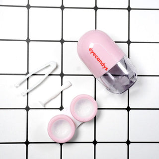 A Pill-Shaped Contact Lens Case Kit on a white and black grid background. The kit includes a pink and transparent contact lens case with the "EyeCandys" logo, soft-tipped tweezers, and a lens applicator stick. The case is opened displaying two compartments for the lenses.