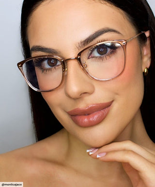 A woman with long dark hair and manicured nails wears EyeCandys Posh Square unisex rectangular glasses, natural makeup, and smiles gently, resting her chin on her hand against a plain light background.