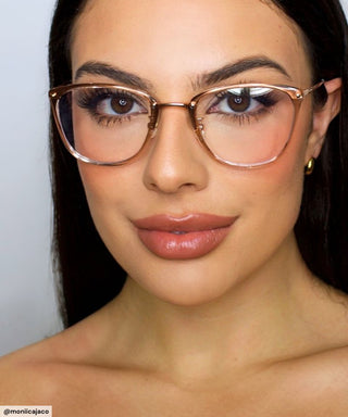 A woman with long dark hair models EyeCandys Posh Square full-frame glasses with large, translucent frames, flawless makeup, bold brows, nude lips, and gold hoops against a plain light background.