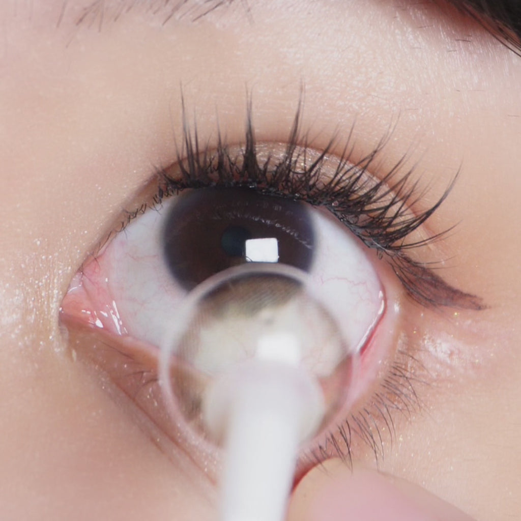 Sequential images demonstrating application of EyeCandys Isla Mirage Grey contact lenses modeled by someone with dark brown eyes and natural eye makeup. The image also includes a close-up of the contact lens.