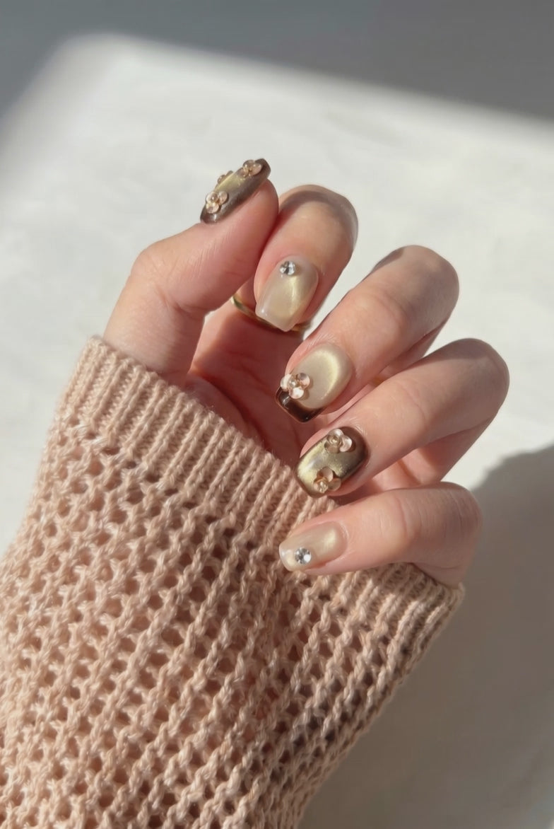 Wearing EyeCandys Gilt Trip Press-On Nails, a glossy gold cat-eye base with bright bands of light, alternating with translucent floral and pearl accents in a short square shape