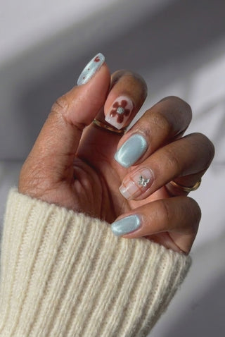 Video alt text: Hand wearing EyeCandys Blue Period press on nails in a short square shape, featuring pale blue cat-eye magnetic nails, sheer floral accent nails with hand-painted brown flowers, blue polka dot nail with a tiny heart, brown grid nail with a silver butterfly charm, softly reflecting light from different angles.
