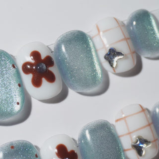 EyeCandys Blue Period Press-On Nails featuring pale blue cat-eye and sheer nails with hand-painted brown flowers, grid illustrations, scattered brown dots, a tiny brown heart, and silver butterfly charm in a short square shape, moody sketched manicure