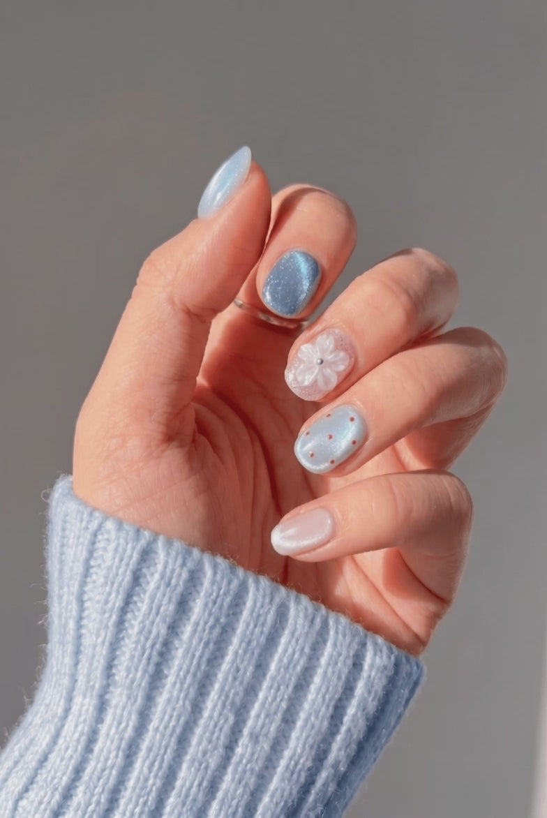 Hand wearing EyeCandys Petal Pusher press on nails in a short round shape, featuring pearlescent blue and steel-blue cat-eye nails, a soft pink nail with a 3D white flower and silver bead center, a grey-blue cat-eye nail with red polka dots, and a blush pink pearlescent chrome accent, shimmering as the hand moves.