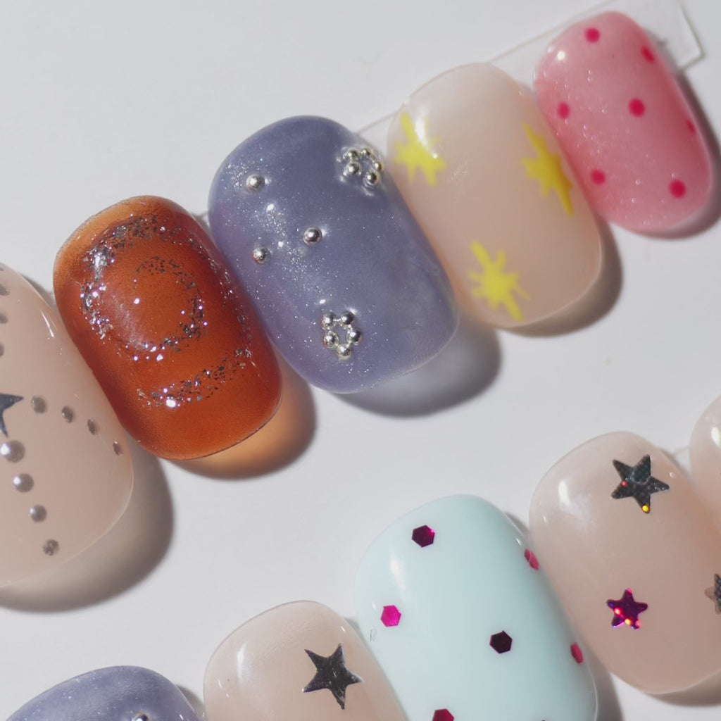 EyeCandys Confetti Crush Press-On Nails featuring nude, amber, and periwinkle cat-eye nails with silver star charms, beads, yellow stars, polka dots, sequin stars, and rainbow confetti glitter in a short squoval shape, trendy colorful party manicure