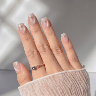 Hand wearing EyeCandys Soft Money press on nails in a short square shape, featuring sheer champagne pearl cat-eye nails with pale pink glitter cat-eye French tips and a tiny crystal accent on each nail, shimmering softly as the hand moves in the light.