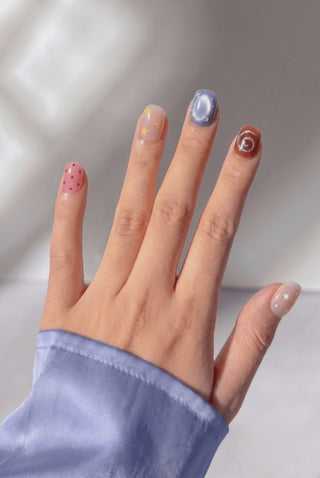 Hand wearing EyeCandys Confetti Crush press on nails in a short squoval shape, featuring warm nude, amber, periwinkle blue cat-eye, and pale mint nails with silver stars, beads, glitter swirls, colorful polka dots, confetti, and rainbow glitter accents, sparkling as the hand moves.