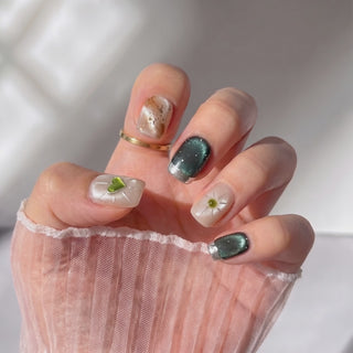 Hand wearing EyeCandys Field Note press on nails in a short square shape, featuring champagne pearl cat-eye nails, a 3D olive-green heart accent with gold sunburst detailing, a brown and rust cat-eye marble nail with fine silver glitter, and deep teal cat-eye nails with silver chrome French tips, reflecting light as the hand moves.