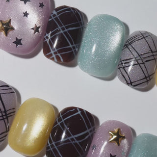 EyeCandys Show and Tell Press-On Nails featuring pale lavender pearl nails with silver stars and gold star charm, chocolate brown plaid, pale blue cat-eye, purple plaid, and butter yellow in a short squoval shape, trendy mixed-design manicure