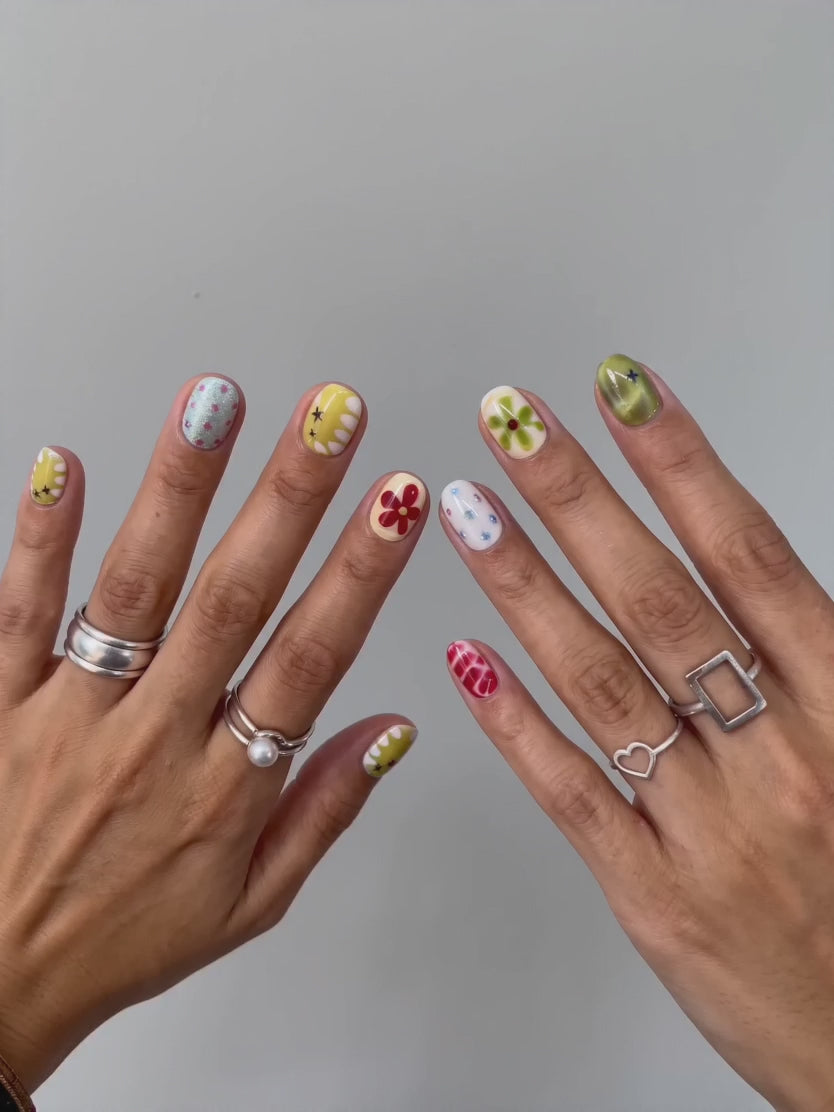 Hand wearing Eyecandys Pucker Up press on nails, featuring citrus-core olive yellow and cream bases with abstract red swirls, floral accents, and holographic details in a short round shape