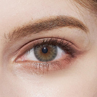 A detailed shot of a model's eye showcasing the Pink Label Flirty Grey contact lens, accompanied by peach-colored eyeshadow