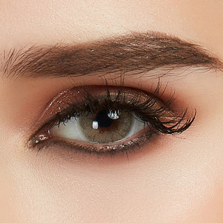 Glossy Ivory hazel color contact lens on top of a dark eye paired with smoky brown eyeshadow and curled wispy eyelashes, above the contact lens design.